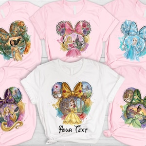 May include: Pink and white t-shirts featuring watercolor designs of Disney princesses within Mickey Mouse ear silhouettes. Each shirt showcases a different princess, including Pocahontas, Aurora, Elsa, Belle, Rapunzel, and Tiana.