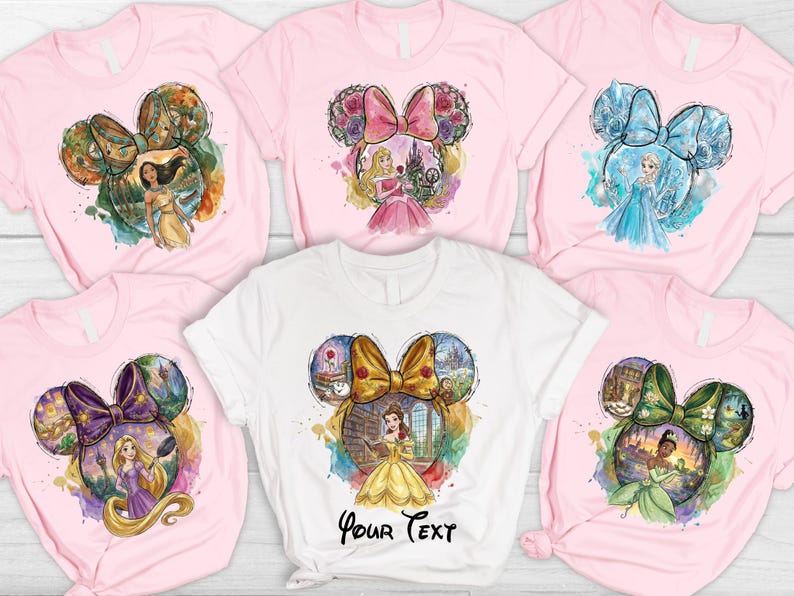 May include: Pink and white t-shirts featuring watercolor designs of Disney princesses within Mickey Mouse ear silhouettes. Each shirt showcases a different princess, including Pocahontas, Aurora, Elsa, Belle, Rapunzel, and Tiana.