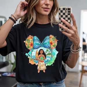 May include: Black t-shirt featuring a colorful graphic of a character from a popular animated film. The design includes a blue bow, floral accents, and a tropical beach scene. The character is holding a glowing orb.