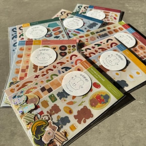May include: A collection of multiple clear plastic sleeves containing various sticker sheets. Each sleeve features a circular white label with a line-art bunny illustration and the text 'oops! sticker pack'. The stickers depict colorful food items, animals, and shapes.