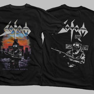 May include: Black t-shirt featuring the band name "Sodom" in white, with a graphic of a figure in a gas mask holding a rifle. The front design includes a cityscape and the text "Persecution Mania". The back has a similar design.