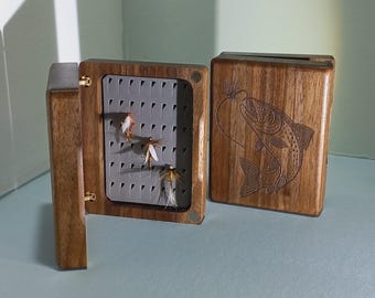 May include: A wooden fly fishing box with a hinged lid and a gray interior. The box is open, revealing several artificial flies. The exterior features a carved fish design on the lid.