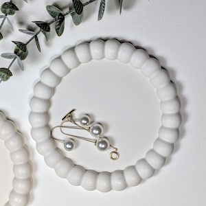 May include: A white, round decorative tray with a beaded edge. A gold-toned ear cuff with faux pearls rests on the tray. The tray is placed on a white surface, with a sprig of greenery in the background.