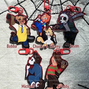 K&ouml;nnte beinhalten: Eine Sammlung von Schl&uuml;sselanh&auml;ngern mit roten R&auml;ndern, die Cartoon-Horrorfiguren zeigen: Bubba, Chucky & Tiffany, Jason, Michael und Freddy. Jeder Schl&uuml;sselanh&auml;nger hat einen roten Verschluss und zeigt eine Figur mit einer Waffe. Die Figuren sind in verschiedenen Posen.