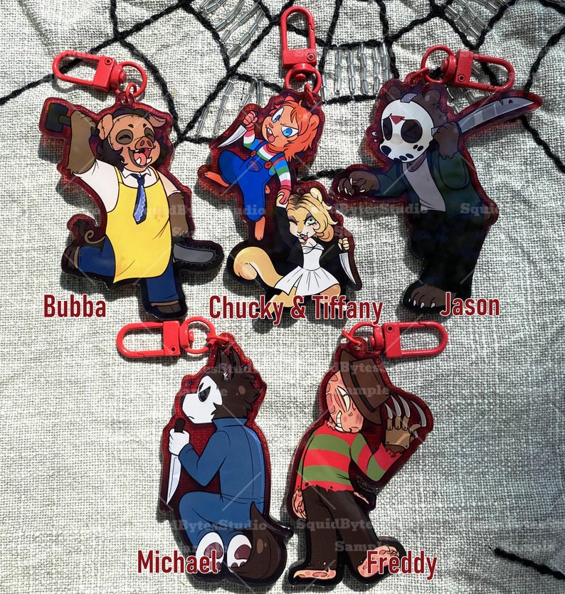 K&ouml;nnte beinhalten: Eine Sammlung von Schl&uuml;sselanh&auml;ngern mit roten R&auml;ndern, die Cartoon-Horrorfiguren zeigen: Bubba, Chucky & Tiffany, Jason, Michael und Freddy. Jeder Schl&uuml;sselanh&auml;nger hat einen roten Verschluss und zeigt eine Figur mit einer Waffe. Die Figuren sind in verschiedenen Posen.