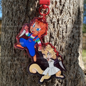 K&ouml;nnte beinhalten: Ein roter Schl&uuml;sselanh&auml;nger mit zwei Acryl-Anh&auml;ngern mit Zeichentrickfiguren. Ein Anh&auml;nger zeigt eine Figur mit orangefarbenem Haar, blauen Augen und einem Messer, die blaue Latzhosen tr&auml;gt. Der andere Anh&auml;nger zeigt eine Figur mit blondem Haar, einem wei&szlig;en Kleid und einem Messer.