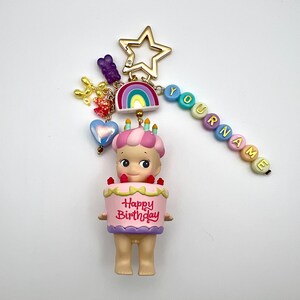 Puede incluir: Un llavero colorido con una figura de beb&eacute; que sostiene un pastel rosa con el texto "Happy Birthday". El llavero incluye una estrella dorada, un arco&iacute;ris y cuentas que deletrean "YOURNAME". Los dijes adicionales incluyen un oso morado, flores doradas y un coraz&oacute;n azul.