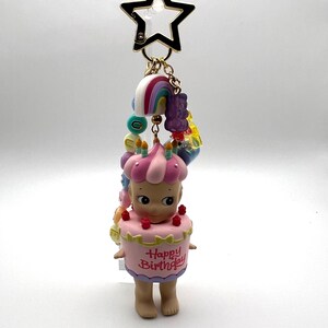 Puede incluir: Un llavero con una figura vestida de tarta de cumplea&ntilde;os rosa, con la inscripci&oacute;n "Happy Birthday". Tiene un cierre dorado en forma de estrella y abalorios de arco&iacute;ris. La figura mide unos 5 cm.