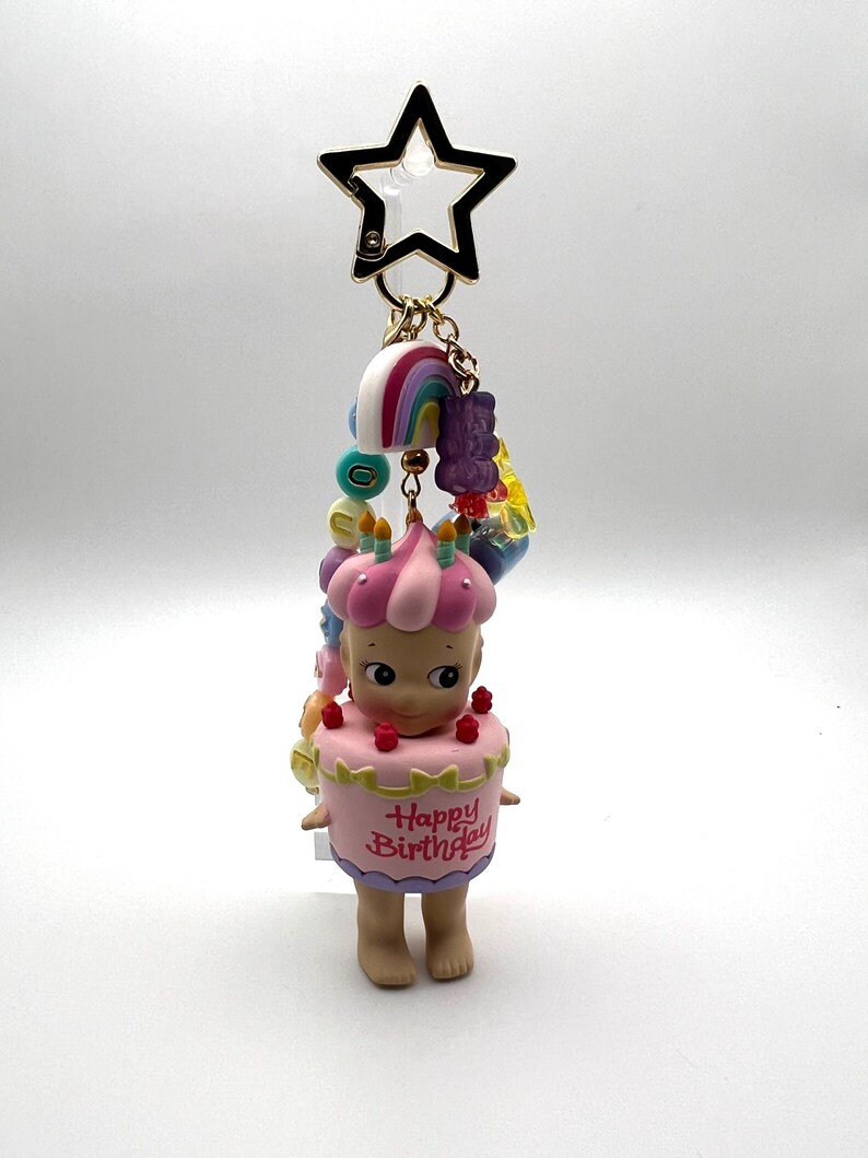 Puede incluir: Un llavero con una figura vestida de tarta de cumplea&ntilde;os rosa, con la inscripci&oacute;n "Happy Birthday". Tiene un cierre dorado en forma de estrella y abalorios de arco&iacute;ris. La figura mide unos 5 cm.