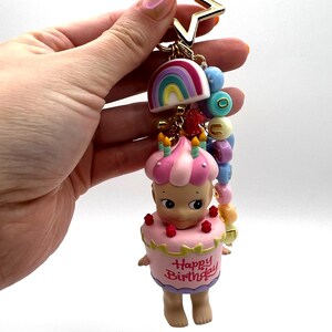 Puede incluir: Un llavero con una estrella dorada, un arco&iacute;ris y una figura vestida de pastel. La figura tiene un dise&ntilde;o de pastel rosa y blanco con las palabras "Happy Birthday". El llavero incluye cuentas de colores.