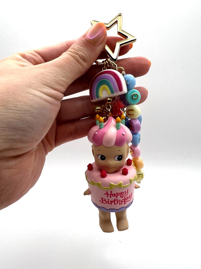 Puede incluir: Un llavero con una estrella dorada, un arco&iacute;ris y una figura vestida de pastel. La figura tiene un dise&ntilde;o de pastel rosa y blanco con las palabras "Happy Birthday". El llavero incluye cuentas de colores.