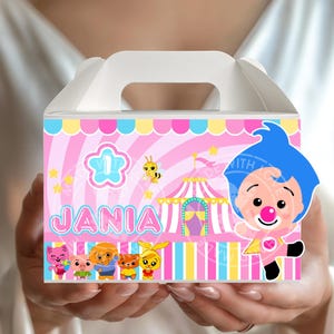 Puede incluir: Una caja de regalo de cartón blanco con asa, decorada con un tema de circo rosa y amarillo. La caja presenta el nombre "JANIA" y personajes de dibujos animados. Un payaso de dibujos animados está en el lado derecho.
