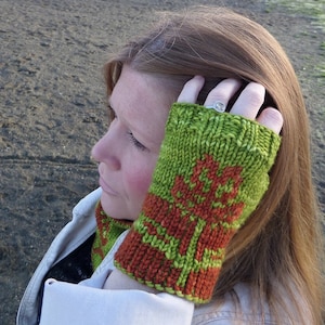 May include: A close-up of a woman's hand wearing a green and orange knitted fingerless glove. The glove has a floral pattern and is made with a chunky yarn.