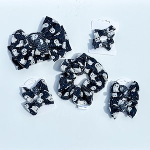 May include: A collection of hair accessories featuring a black fabric with a white ghost pattern. The set includes one large knotted headband, one large fabric scrunchie, and three pairs of small hair bows attached to white display cards.