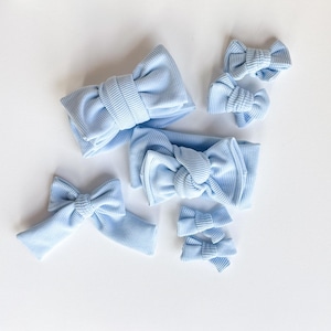 May include: A collection of light blue ribbed fabric bows and headbands. The set includes various sizes of bows and a wide headband, all in a soft, pastel blue color. The bows are arranged on a white surface.
