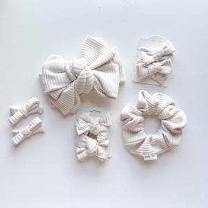May include: A collection of cream-colored hair accessories made from ribbed fabric. The set includes one large knotted bow headband, two pairs of small bow hair clips on white cards, and one large fabric scrunchie with a small white brand tag.