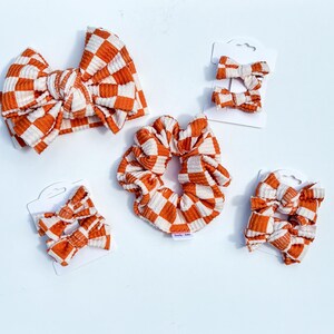May include: A collection of hair accessories featuring a burnt orange and white checkered pattern on a ribbed fabric. The set includes one large knotted headband, one large scrunchie, and four small hair bows attached to white display cards.