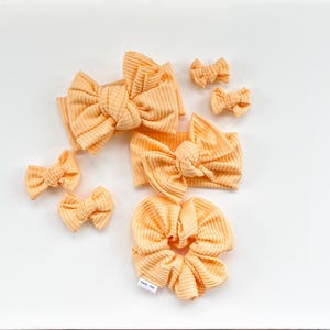 May include: A collection of peach-colored hair accessories, including headbands, bows, and a scrunchie. The items are made of a ribbed fabric and are arranged on a white surface. The bows vary in size, and the scrunchie has a small tag attached.