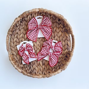 May include: A collection of hair bows featuring a red and white gingham check pattern. The bows are attached to white display cards and include one large bow, two smaller bows on one card, and two medium bows on another card, all with red trim.