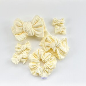 May include: An assortment of cream-colored corduroy hair accessories, including several bow-shaped hair clips, a scrunchie, and a headband. The accessories are arranged on a white surface, showcasing their textured fabric and design.