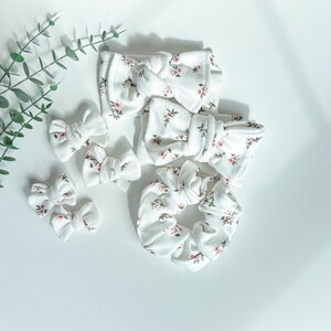 May include: A collection of white fabric hair accessories with a delicate floral pattern in pink and green. The set includes several bow-shaped hair clips, headbands, and a scrunchie, all arranged on a white surface.