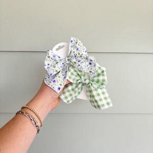 May include: Two fabric hair bows. One bow features a white background with purple floral print. The other bow has a green and white gingham pattern. Both bows are attached to white cardboard.