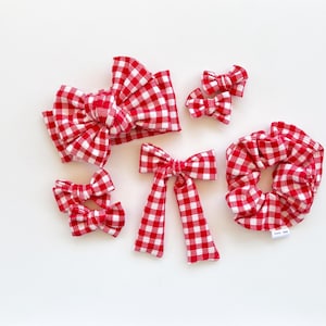 May include: A collection of red and white gingham hair accessories including a large knotted headband, a scrunchie with a small white tag, a bow with two long hanging tails, and four smaller hair bows of varying sizes arranged on a plain surface.