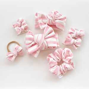 May include: A collection of pink and white striped hair accessories, including headbands, scrunchies, and bow clips. The items are arranged on a white surface, showcasing the various styles and sizes available. The accessories are made of a ribbed fabric.