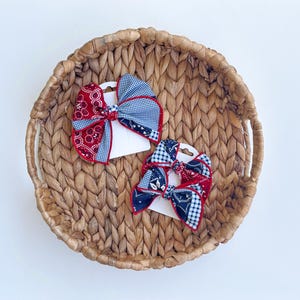 May include: Two hair bows featuring a patchwork design of red and navy blue bandana patterns mixed with black and white gingham checks. Each bow is secured to a white card backing, showcasing a multi-layered fabric construction with red trim.