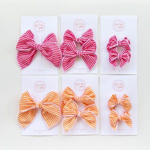 May include: Six fabric hair bows in various sizes, arranged on white cards. The bows are striped in pink and white, and orange and white. The bows are tied in the center and have a classic bow shape. The cards have a logo in the upper left corner.