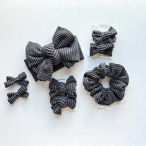 May include: A collection of hair accessories in a dark gray and black ribbed striped fabric, including one large knotted headband, one scrunchie, and four small hair bows attached to white display cards, featuring the text Truly Jane.