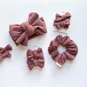 May include: A set of hair accessories in a dusty rose and white striped ribbed fabric. The collection includes one large knotted headband, one circular scrunchie, and four small bow clips attached to white display cards for organization.