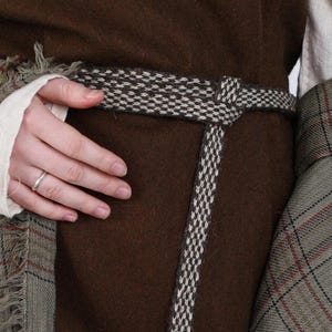 May include: A brown dress with a woven belt and a fringed plaid shawl. The belt is black and white, and the shawl has red and green accents. The dress is a solid color, and the shawl is draped over the shoulders.