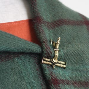 May include: A brass-toned metal brooch with a mechanical, T-shaped design featuring coiled springs, secured to a green and maroon plaid wool fabric garment that is layered over a plain orange fabric and a white fabric collar.