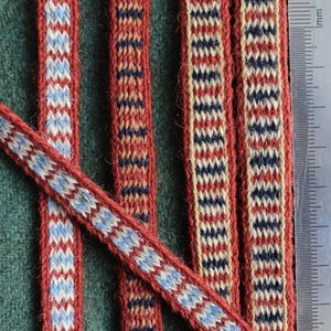 May include: Four strips of woven fabric trim featuring geometric patterns in red, blue, cream, and light blue. A metal ruler is placed alongside the strips, showing a scale in centimeters, with the longest visible strip measuring 13 centimeters.