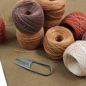 May include: A collection of round yarn balls in various shades of brown, tan, rust, and cream, arranged in a cluster. A pair of metal thread snips with a simple U-shaped handle is positioned to the left of the yarn balls on the surface.
