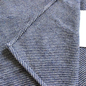 K&ouml;nnte beinhalten: Nahaufnahme eines blau-wei&szlig; gestreiften Stoffes mit einer aufgen&auml;hten Tasche und einem leeren wei&szlig;en Etikett. Der Stoff hat ein strukturiertes Aussehen und ein diagonales Muster. Die Tasche ist mit einer Kontrastnaht gen&auml;ht.