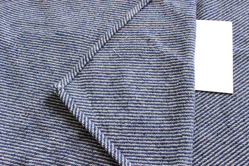 K&ouml;nnte beinhalten: Nahaufnahme eines blau-wei&szlig; gestreiften Stoffes mit einer aufgen&auml;hten Tasche und einem leeren wei&szlig;en Etikett. Der Stoff hat ein strukturiertes Aussehen und ein diagonales Muster. Die Tasche ist mit einer Kontrastnaht gen&auml;ht.