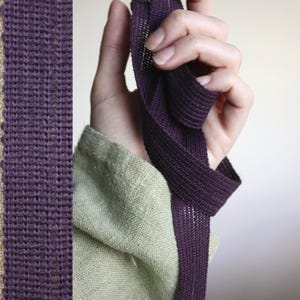 May include: A length of dark purple woven ribbon held in a hand, with a section of the ribbon displayed vertically on the left. The ribbon has a textured, open weave. A section of tan fabric is visible on the left edge.
