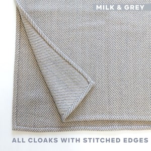 K&ouml;nnte beinhalten: Nahaufnahme eines gefalteten, hellgrauen Umhangs mit gen&auml;hten Kanten. Der Umhang hat ein Fischgr&auml;tenmuster. Der Text "MILK & GREY" steht oben. Der Text "ALL CLOAKS WITH STITCHED EDGES" steht unten.