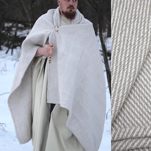 May include: A light gray and white striped cloak with a gold-colored clasp. The cloak is part of a medieval-style outfit, with a long, cream-colored tunic and a beaded accessory. The outfit is worn outdoors in a snowy environment.