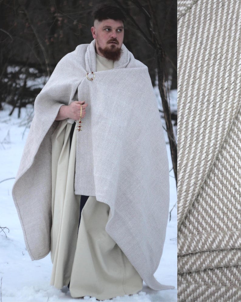 K&ouml;nnte beinhalten: Ein hellgrauer und wei&szlig; gestreifter Umhang mit einem goldfarbenen Verschluss. Der Umhang ist Teil eines Outfits im mittelalterlichen Stil, mit einer langen, cremefarbenen Tunika und einem Perlenaccessoire. Das Outfit wird im Freien in einer verschneiten Umgebung getragen.