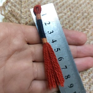 Pode incluir: Um close-up de um tassel vermelho com detalhes pretos e laranja. O tassel mede aproximadamente 10 cm de comprimento e est&aacute; sendo segurado em uma m&atilde;o ao lado de uma r&eacute;gua.