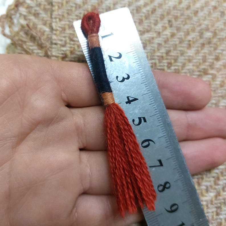 Pode incluir: Um close-up de um tassel vermelho com detalhes pretos e laranja. O tassel mede aproximadamente 10 cm de comprimento e est&aacute; sendo segurado em uma m&atilde;o ao lado de uma r&eacute;gua.