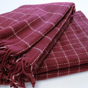 Pu&ograve; includere: Una coperta in plaid bordeaux con frange bianche. La coperta &egrave; piegata a met&agrave; e ha una texture morbida e soffice.