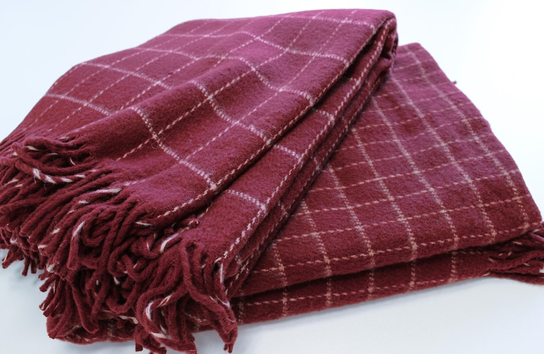 Pu&ograve; includere: Una coperta in plaid bordeaux con frange bianche. La coperta &egrave; piegata a met&agrave; e ha una texture morbida e soffice.
