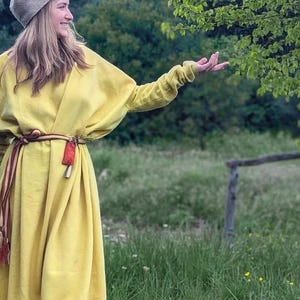 Puede incluir: Un vestido amarillo de manga larga con corte holgado, combinado con un gorro de punto beige y un cinturón de tela multicolor con rayas rojas y marrones. El vestido tiene un cuello redondo sencillo, llega hasta los tobillos y se ajusta a la cintura.