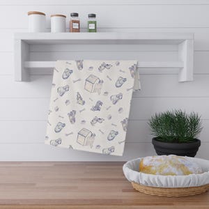 May include: A white kitchen towel with a repeating pattern of blue dog illustrations, dog houses, and bones. The towel is hanging on a white shelf above a wooden countertop. Other items on the shelf include jars and spices.