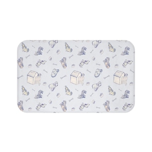 Puede incluir: Una alfombra de baño rectangular de color azul claro con un patrón repetido de perros ilustrados, casetas para perros, huesos y cuencos de comida. Los perros están en varias poses, y las ilustraciones son en tonos marrones, grises y amarillos.