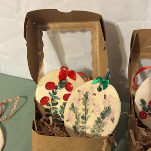 Op de afbeelding: Ovale keramische ornamenten met handgeschilderde bloemmotieven, gepresenteerd in een bruine kartonnen doos met een helder venster. Eén ornament heeft rode bessen, de andere, roze bloemen. Elk heeft een lint om op te hangen.